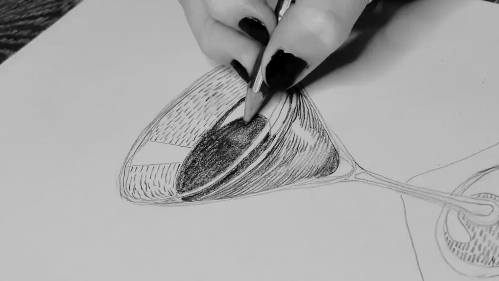 Black and white close up video of a woman’s hand with black nails holding a pencil drawing a martini glass with black liquid in it on white paper.